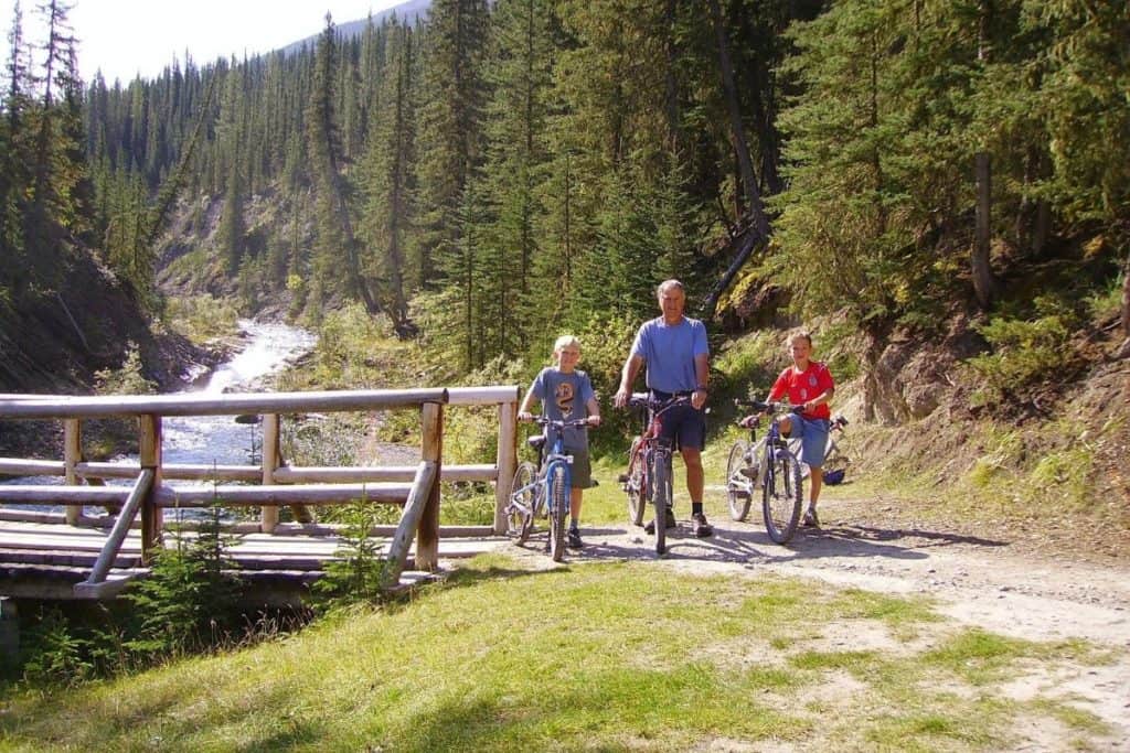Dos niños y el abuelo recorren en bicicleta el sendero del arroyo Goat cerca de un río en Canmore. Deportes más populares en Canadá.